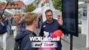 Ein Mitarbeiter des WDR erklärt einer Besucherin des Mettwurstmarkts in Ennigerloh die neue WDR aktuell-App auf einem Bildschirm