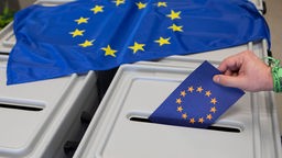 Man sieht eine Hand, die einen Umschlag in die Wahlurne wirft, darauf liegt eine Europaflagge