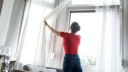 Eine Frau zieht in einer Wohnung eine Gardine vor einem gekippten Fenster auf