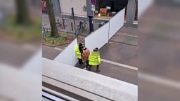 Handyaufnahme, auf der zwei Sicherheitsmänner zu sehen sind und ein Jeck. Sie stehen an einer Straßenabsperrung, die das Gebiet rund um den Zülpicher Platz abschirmt.