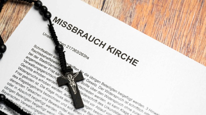 Ein Holzkreuz liegt auf einem Schriftstück mit der Überschrift "Missbrauch Kirche"