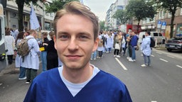 Ein junger Mann mit weißem T-Shirt und blauem Arztkittel, im Hintergrund viele Menschen mit Plakaten und Transparenten