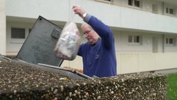 Jürgen Kaminski schmeißt eine weiße Mülltüte draußen in einen Mülleimer