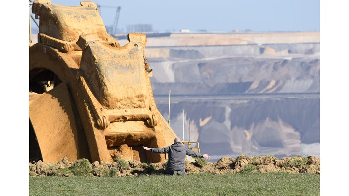 Ein Mann sitze vor einem Schauffelradbagger, hat die Arme weit ausgebreitet, im Hintergrund ist ein Braunkohletagebau zu sehen