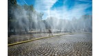 Ein Wasserschlauch liegt auf Kopfsteinpflaster, aus zahlreichen Löchern sprüht feiner Wassernebel, ein Kind springt fröhlich über den Schlauch