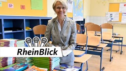 NRW-Schulministerin Dorothe Feller (CDU) sitzt in einem leeren Kalssenraum und hat hinter einen Stapel Schulbücher ein Buch aufgeschlagen