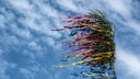 Ein Maibaum weht im Wind mit vielen bunten Bändern, im Hintergrund Wolken.