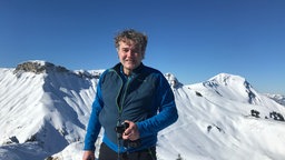 Georg Bayerle mit BR-Mikrofon an einem sonnigen Tag in schneebedeckten Bergen.