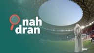 Mann mit traditioneller Kleidung steht in Fuball-Stadion in Katar. Daneben das Podcast-Logo: Schriftzug "nah dran", daneben eine kleine Lupe.