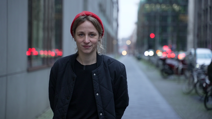 Das Bild zeigt die Reporterin Helena Daahler mit roter Mütze, schwarzer Jacke und schwarzem Pulli auf einem Gehweg. Im Hintergrund sieht man unscharf eine Straße. 