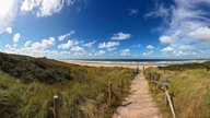 Ein Weg der zum Strand an der Nordseeküste führt. An den Seiten befinden sich Wiesen und Hügel, die Sonne scheint und es befinden sich ein paar Wolken im blauen Himmel.