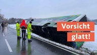 Ein Linienbus liegt auf der Seite im Straßengraben; es liegt Schnee; im Hintergrund sieht man ein Feuerwehrauto; neben dem Bus stehen Männer in Neonkleidung; auf rotem Banner "Vorsicht glatt!"