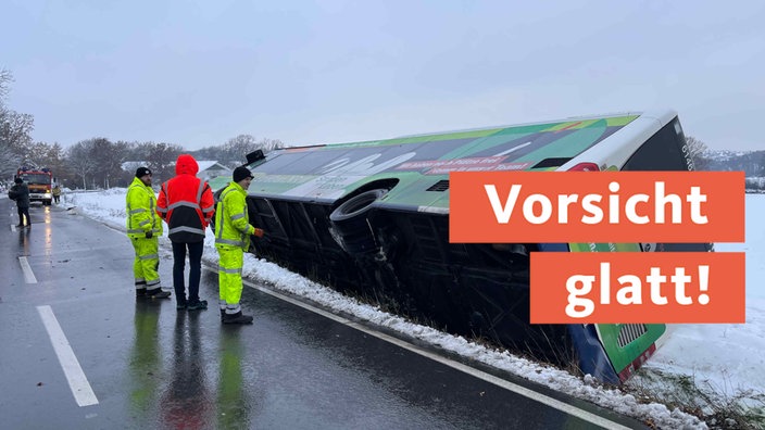 Ein Linienbus liegt auf der Seite im Straßengraben; es liegt Schnee; im Hintergrund sieht man ein Feuerwehrauto; neben dem Bus stehen Männer in Neonkleidung; auf rotem Banner "Vorsicht glatt!"