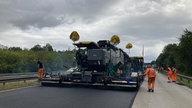 Ein Kompaktasphaltgerät teert die Fahrbahn, daneben Arbeiter in orangener Arbeitskleidung