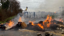 Quer über eine Straße verteilt liegen brennende Strohballen, Paletten und Ziegelsteine. Im Hintergrund mehrere Personen mit weißen Ganzkörperanzügen.