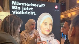 Frauen aus verschiedenen Kulturen stehen vor einem Plakat mit der Aufschrift "Männerberuf ? Bis jetzt vielleicht"