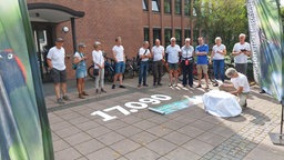 Einige Menschen in weißen T-Shirts stehen in einem Kreis vor dem Kreishaus Kleve. Auf dem Asphalt steht eine Zahl: 17.090