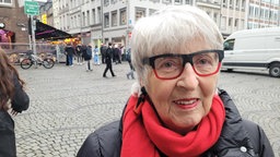 Eine ältere Frau mit weißen Haaren, rotem Schal, schwarzer Jacke und markanter schwarz-roter Brille