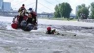 Auf dem Foto ist ein Mann mit rotem Helm, der einen auf dem Wasser liegenden Mann mit sich zieht. Im Hintergrund ist ein Feuerwehrboot.