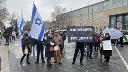 Demonstrierende mit Israel-Flaggen stehen vor der Universtität. Im Hintergrund steht ein Polizeiauto. Auf einem Plakat steht "Nie wieder ist jetzt!"