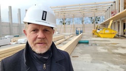 Ein Mann mit weißem Helm und dunkler Jacke und Baustelle im Hintergrund