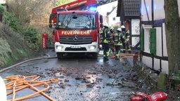 Feuerwehrleute stehen neben einem Feuerwehrauto an der Seite des Fachwerkhauses, auf dem Boden liegt Schutt neben Schläuchen und leeren Feuerlöschern