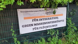 Ein weißes Banner mit großen schwarzen Buchstaben hängt an einem Zaun