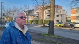 Ein Mann mit grauen Haaren, Jeansjacke und getönter Brille an einer Straße