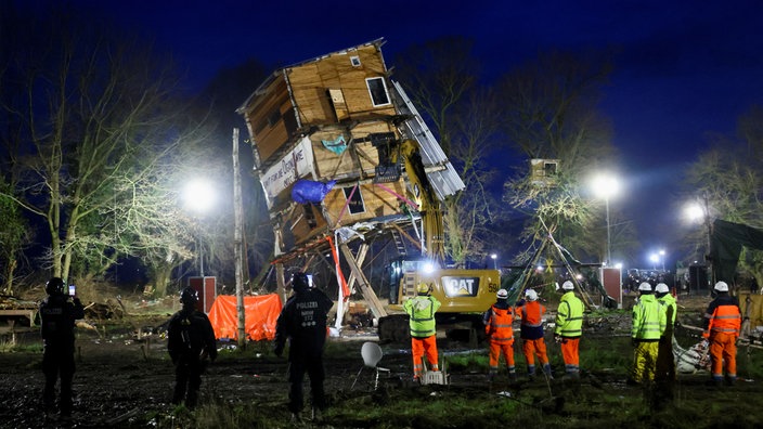 Im Flutlicht: Eine quadratische, circa 10 Meter hohe, mehrstöckige Holzhütte, die mit Bannern behangen ist, befindet sich in Schieflage. Ein gelber Bagger greift nach ihr.