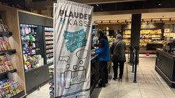 In einem Supermarkt in Kempen hat eine neue "Plauderkasse" eröffnet. Zu sehen ist die Kasse mit einer Beach-Flag davor, auf der "Plauderkasse, hier haben sie alle Zeit der Welt" steht.