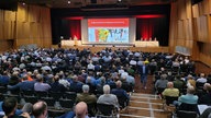 Eine große Halle mit Bühne und ein großen Leinwand, im Saal sitzen viele Männer