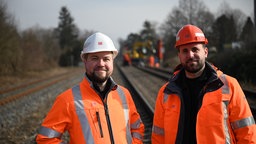 Zwei Männer stehen im Gleisbett eoner Bahnstrecke. Sie schauen in die Kamera. beide tagen orangene Warnjacken. Der linke trägt einen weißen Helm, er grinst und trägt einen Drei-tage-Bart. Der rechte Mann trägt einen roten Helm und ebenfalls Bart.