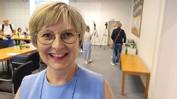 Eine Frau mit Brille und blauem Kleid lächelt, im Hintergrund mehrere Menschen