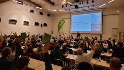 Menschen in Tischreihen halten grüne Kärtchen hoch