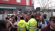 Zwei Ordner stehen mit einer gelben Security-Jacke vor einer Absperrung auf der Zülpicher Straße, an der sich viele Menschen drängen