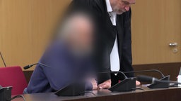 Auf dem Foto ist ein bärtiger Mann im blauen Pullover. Sein Gesicht ist verpixelt. Neben ihm steht sein Anwalt.