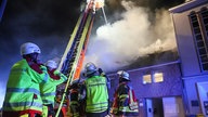 Auf dem Foto sind Feuerwehrleute, die vor einer Drehleiter stehen. Von der Drehleiter aus wird ein stark rauchendes Dach gelöscht.