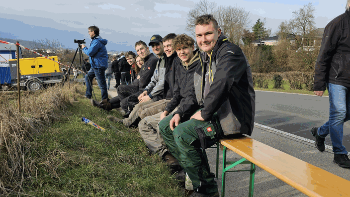 Man sieht sieben Personen der jungen Landwirte auf einer Bierbank sitzen, im Hintergrund hat jemand eine Fotokamera auf einem Stativ befestigt