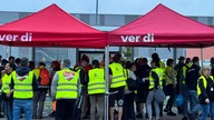 Viele Mitarbeiter mit gelben Warnwesten auf dem Bürgersteig unter zwei roten Pavillons