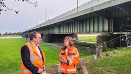 Ein Frau und ein Mann in orangen Warnwesten stehen vor einer Brücke