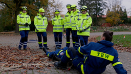 THW Mitarbeiterin liegt auf dem Boden und zeigt einer Gruppe von Kollegen in neongelben Jacken den richtigen Umgang mit verletzten Personen