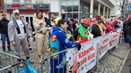 Auf dem Foto sind kostümierte Menschen, die hinter einer Absperrung einem Karnevalsumzug zujubeln. Einer von ihnen hält ein Mikrofon in der Hand.