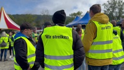 Im Vordergrund drei Männer von hinten mit Warnwesten. Auf einer steht: Wir streiken.