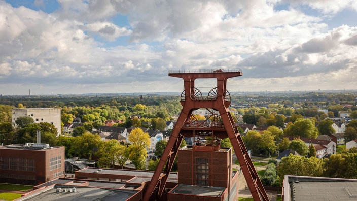 Ein großer roter Förderturm ragt in den Himmel. Im Hintergrund stehen grüne Bäume.