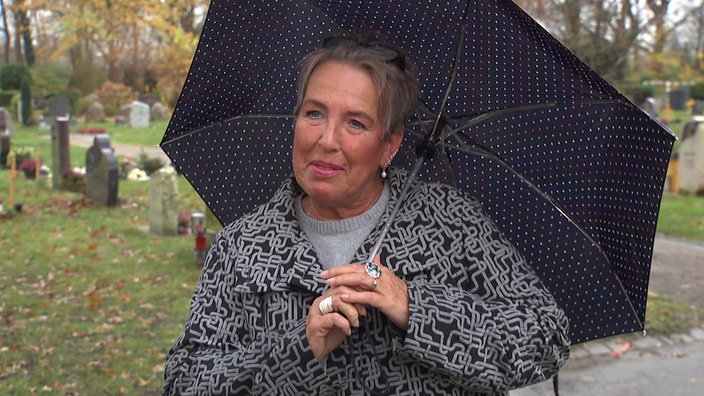 Eine Frau mit einem Regenschirm, in Hintergrund sind Grabsteine zu sehen