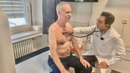 Der Patient Helmut Hoppe sitzt im Behandlungsraum, der Arzt Dr. Messiha hält ein Stethoskop an seine Brust.