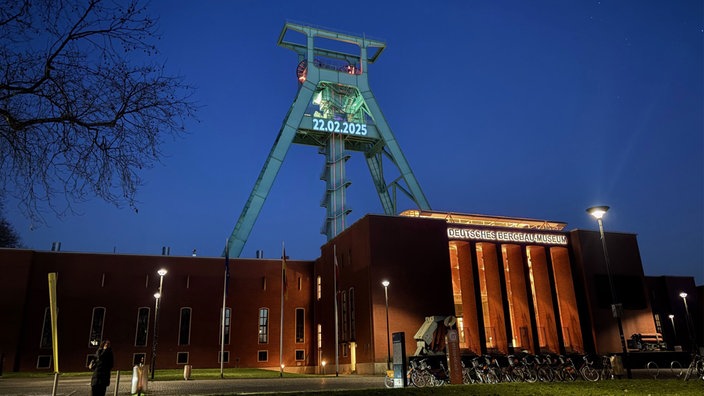 Ein großes Backsteingebäude mit der Aufschrift "Deutsches Bergbau-Museum" in der Dunkelheit. Dahinter ein Turm mit dem Datum 22.02.2025.