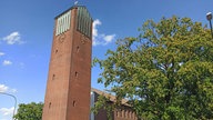 Auf dem Foto ist eine braun gemauerte Kirche mit einer Uhr am Kirchturm und einem goldenen Hahn auf dem Dach.