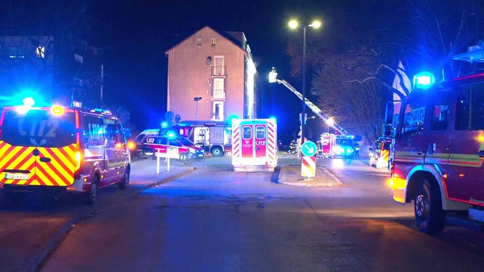 Mehrer Fahrzeuge der Feuerwehr stehen vor einem Haus. Eine Drehleiter wird an dieses haus rangefahren. 