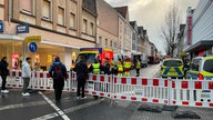 Auf dem Foto ist eine Straße, die abgesperrt ist. Hinter der Absperrung stehen Feuerwehrleute und Polizisten.